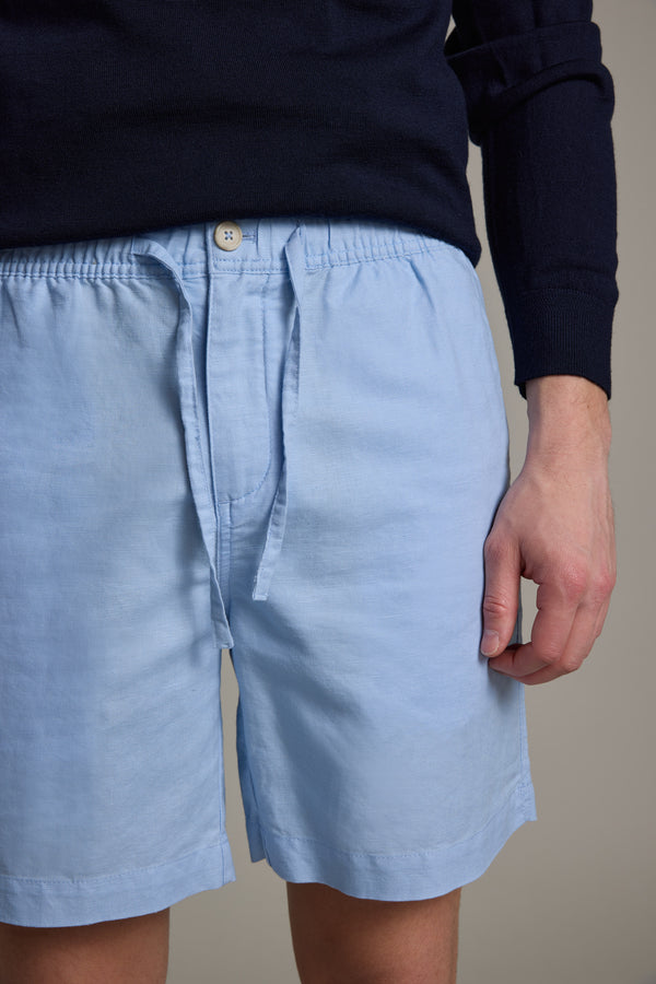 A person models Barkers Jasper Linen Weekend Shorts in light blue and a dark navy long-sleeve top, standing with one arm relaxed at their side. The image is cropped from the shoulder to above the knees.