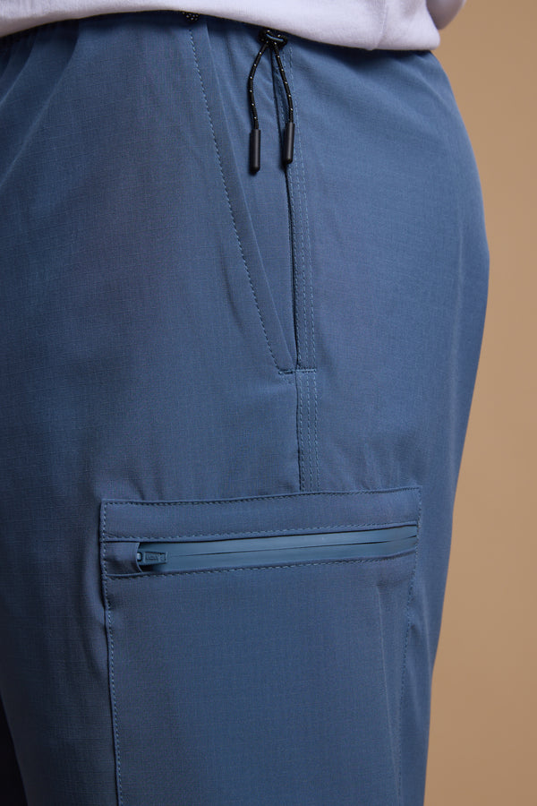 Close-up of a person wearing Legasea Weekend Short by Legasea, featuring blue four-way stretch fabric, zippered pocket, and an elastic drawstring waistband. Shown waist to mid-thigh against a plain beige background.