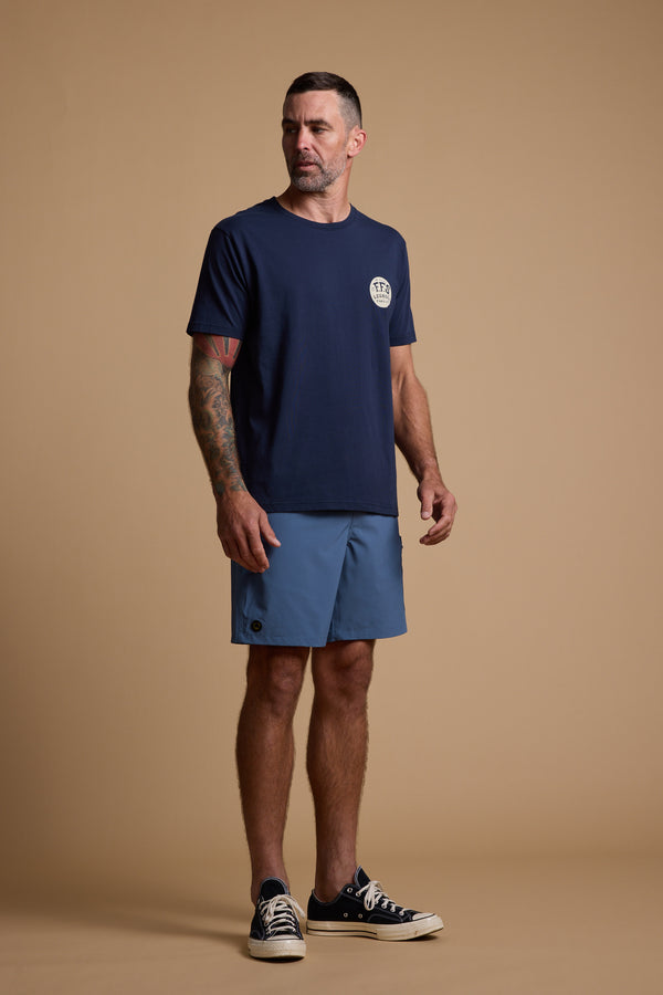 A man with short hair and arm tattoos wears a navy T-shirt, Legasea Weekend Short by Legasea, and black sneakers while standing against a plain beige background.