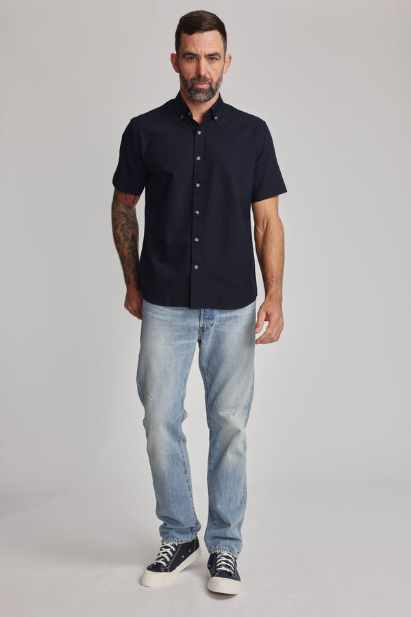 A man with short dark hair and a beard wears the Barkers Archer Oxford Shirt in navy, paired with light blue jeans and black sneakers, standing facing forward against a plain light gray background.