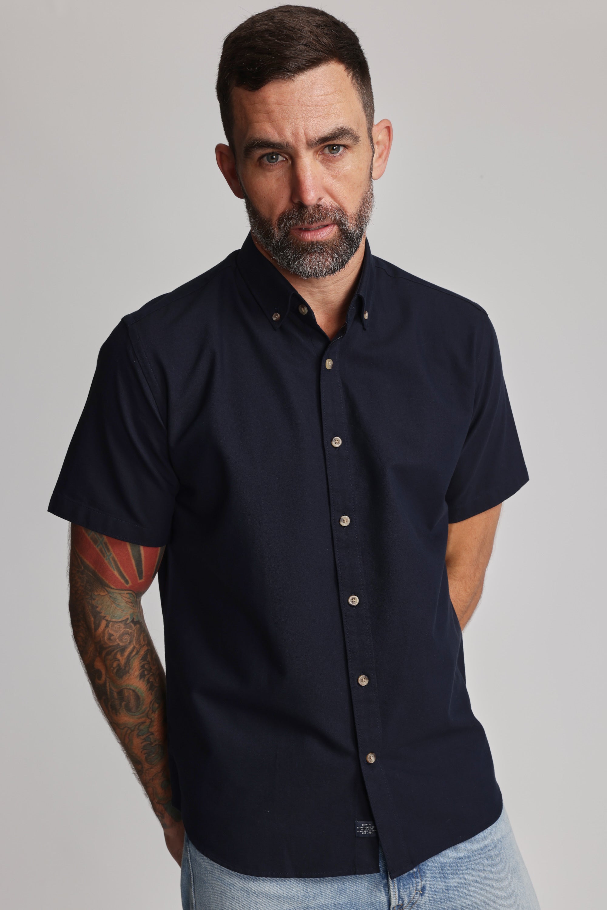 A man with short dark hair, a beard, and a tattooed right arm wears the Barkers Archer Oxford Shirt in navy with a button-down collar and light blue jeans, standing against a plain light background.