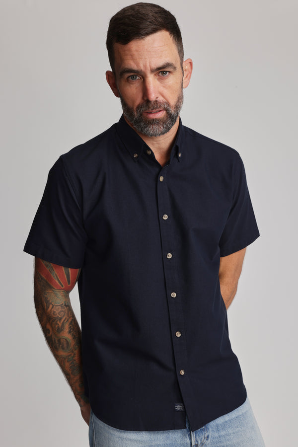 A man with short dark hair, a beard, and a tattooed right arm wears the Barkers Archer Oxford Shirt in navy with a button-down collar and light blue jeans, standing against a plain light background.