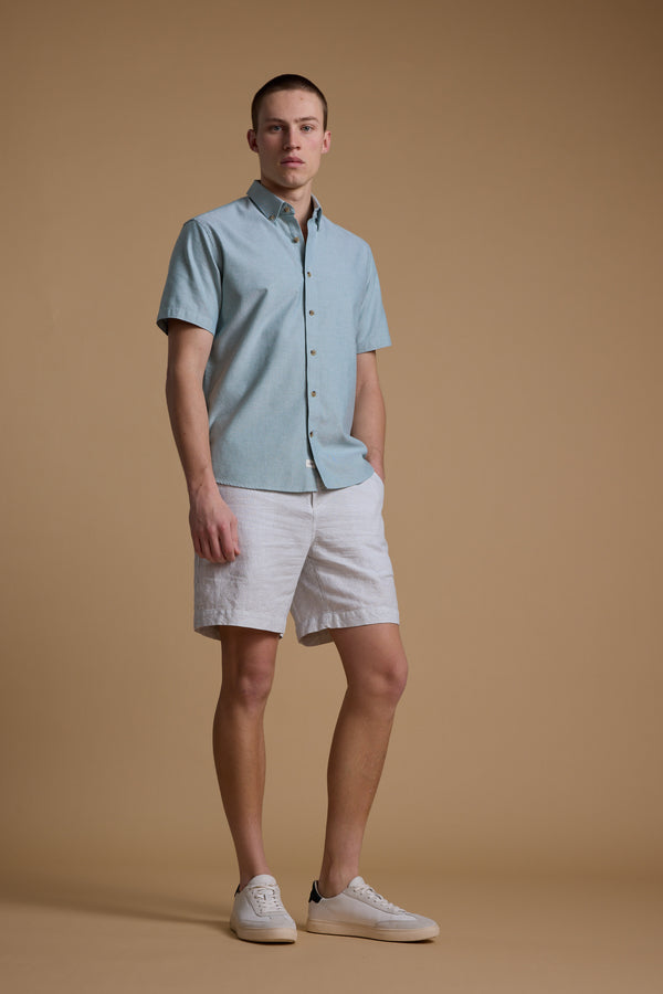 A young man stands against a beige background wearing the Barkers Archer Oxford Shirt in light blue, paired with white shorts and sneakers. He has one hand in his pocket and looks directly at the camera.
