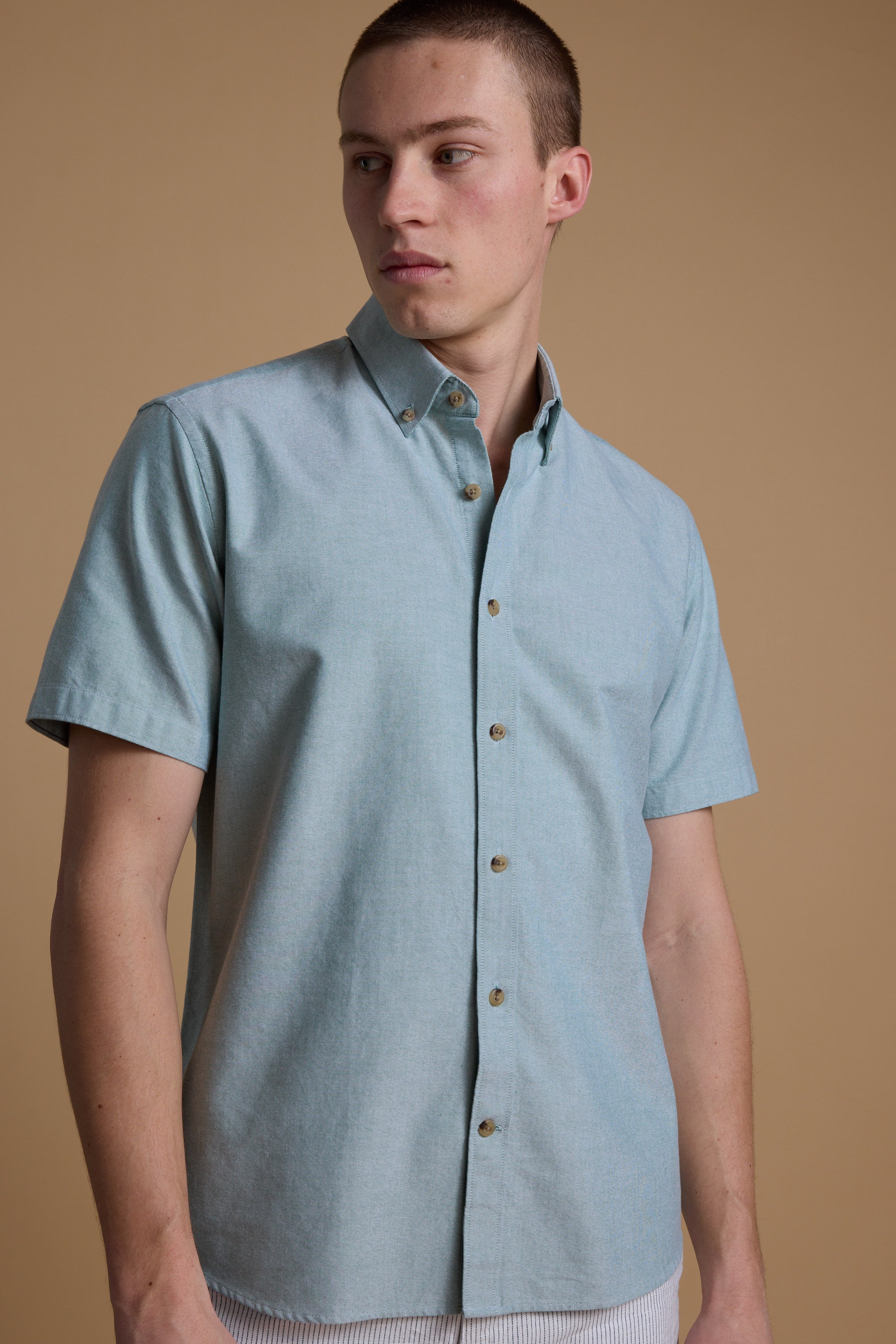 A person in the Barkers Archer Oxford Shirt, featuring a light blue hue and button-down collar, stands against a plain beige background, gazing to the side with a neutral expression.