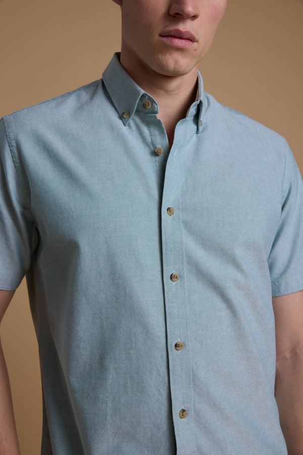 A person models the Barkers Archer Oxford Shirt in light blue 100% cotton with a button-down collar, standing against a plain beige background; their face is partially out of frame.