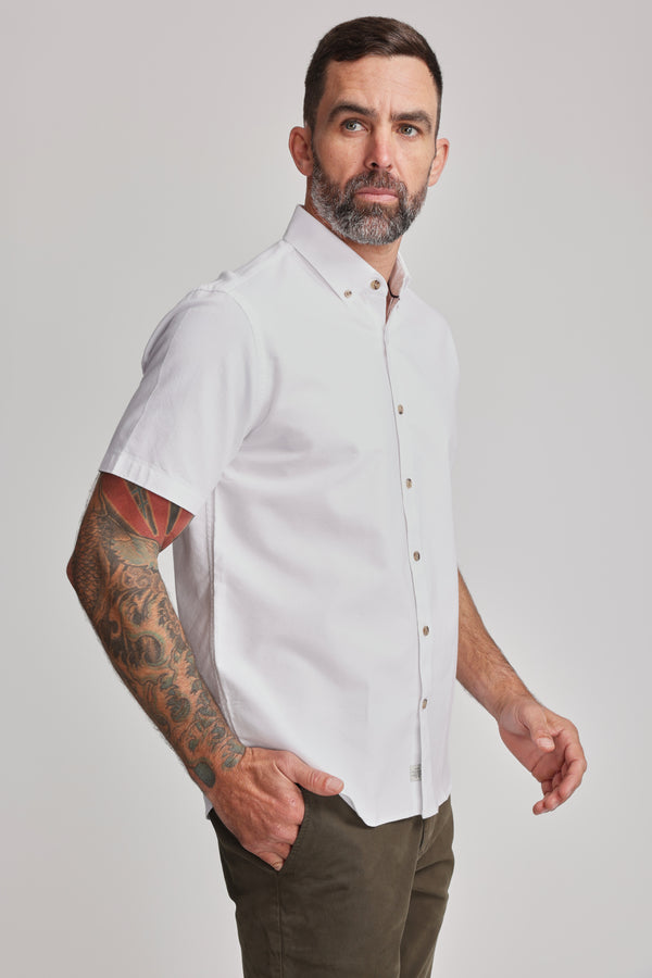 A bearded man with a tattooed right arm wears the Barkers Archer Oxford Shirt—a white, short-sleeve, 100% cotton button-down—paired with olive green pants, standing with one hand in his pocket against a plain light background.
