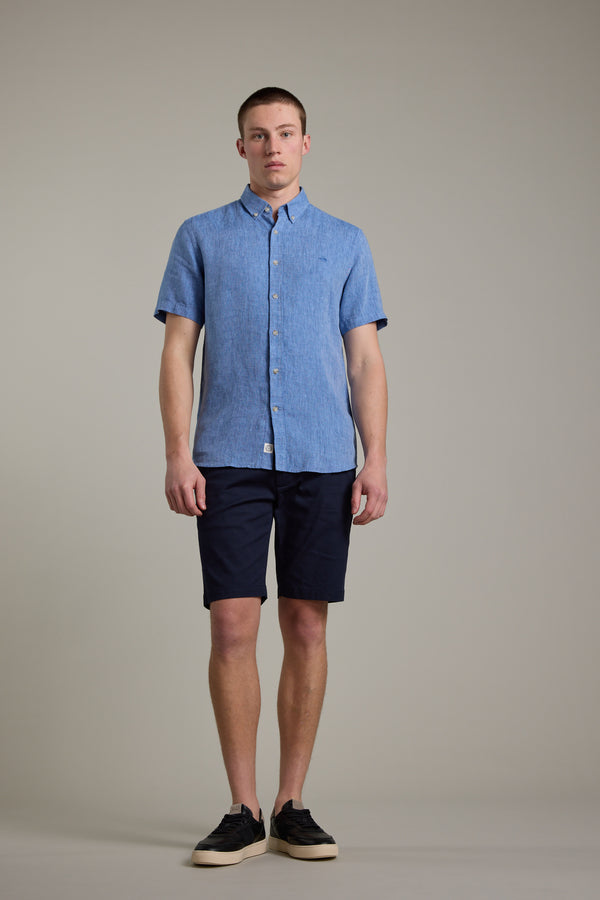 A young man stands indoors against a plain background, wearing the Barkers Gentry Linen Shirt in light blue, paired with navy shorts and black sneakers. He faces forward with a neutral expression.