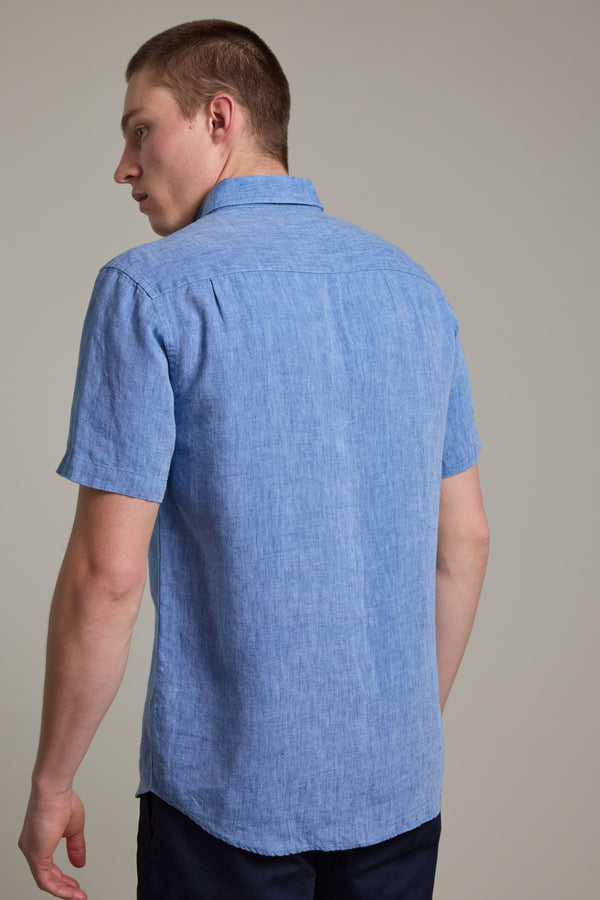 A man with short brown hair is seen from behind, wearing the Barkers Gentry Linen Shirt in light blue and dark pants, standing against a plain light gray background.