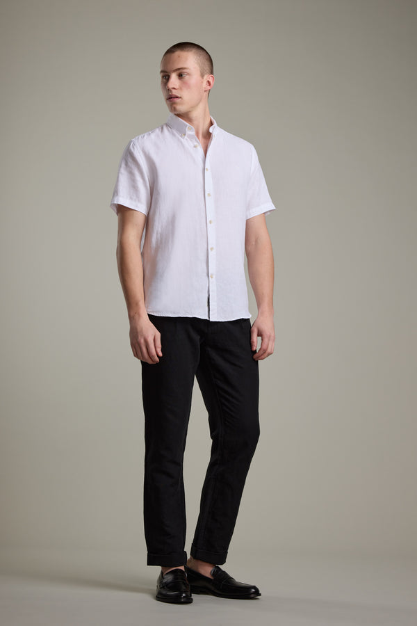 A man stands against a neutral background wearing the Barkers Gentry Linen Shirt in white with subtle crayfish embroidery, paired with black pants and dress shoes, looking slightly to the side with a relaxed posture.