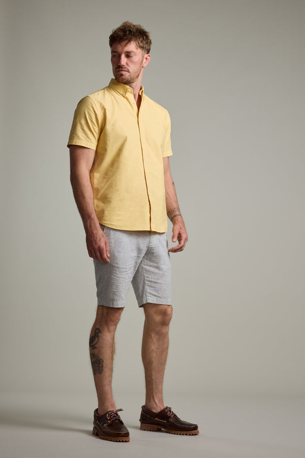 A man with short hair, facial hair, and leg tattoos stands against a plain background in a classic fit Barkers Parker Oxford Shirt in yellow, light gray shorts, and brown boat shoes. The shirt is crafted from responsibly sourced cotton.