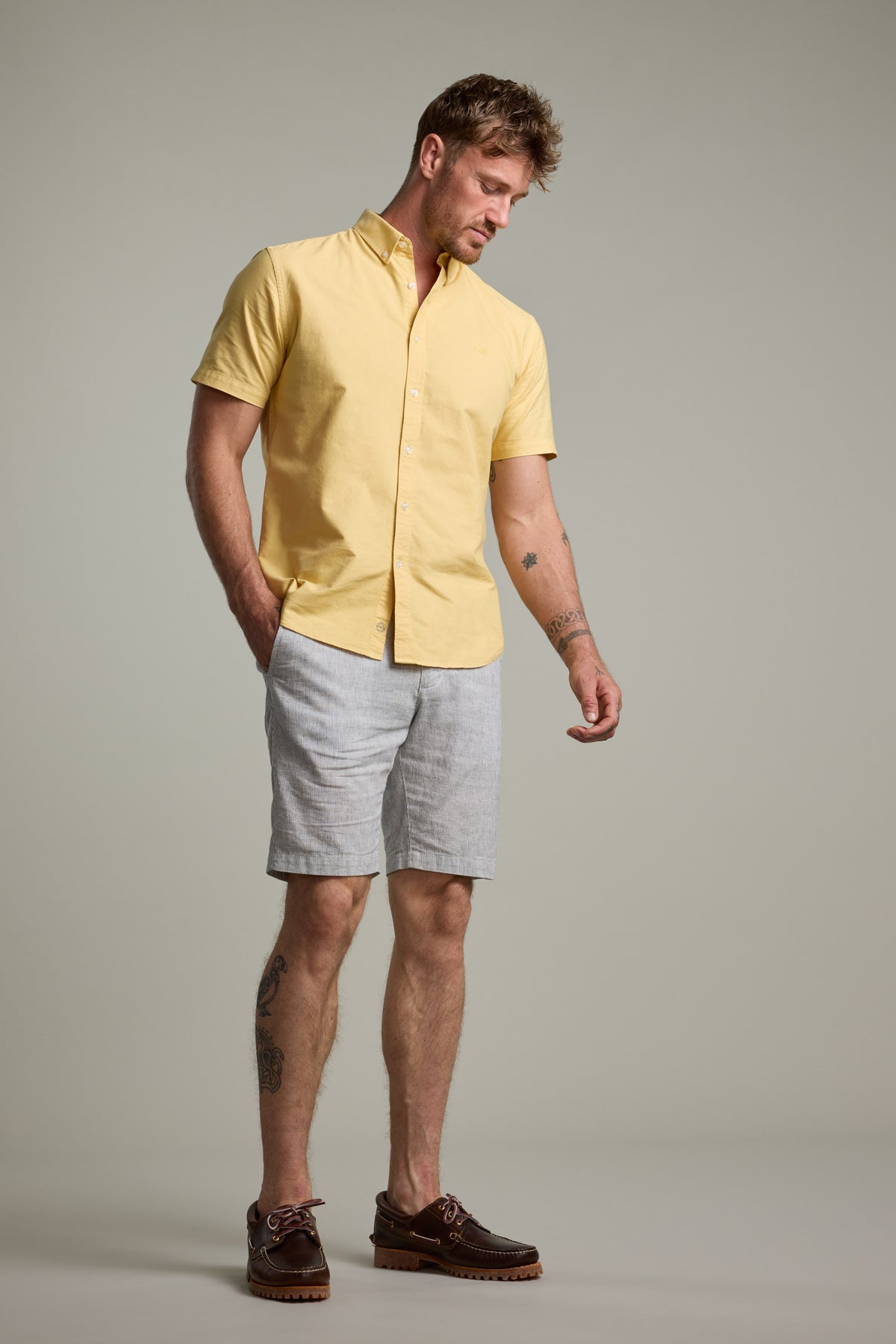 A man stands against a plain background wearing the Barkers Parker Oxford Shirt in classic fit, paired with light gray shorts and brown boat shoes. He has one hand in his pocket and looks down in a relaxed posture.