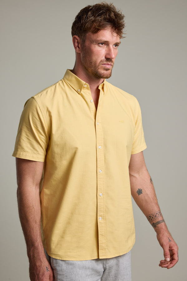 A man with short brown hair and facial hair wears a light yellow Parker Oxford Shirt in classic fit by Barkers, made from responsibly sourced cotton, with light gray pants. He has tattoos on his arms and stands against a plain, light background.