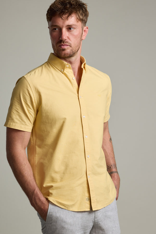 A man with short brown hair and facial hair wears a classic fit Parker Oxford Shirt in light yellow from Barkers, made with responsibly sourced cotton, and light gray pants. He stands with his hands in his pockets against a plain background.