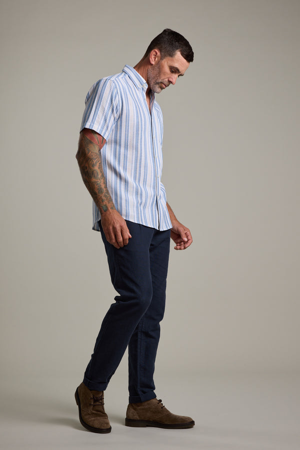 A man with short dark hair and tattooed left arm looks down, wearing the Barkers Merten Stripe Shirt with dark blue pants and brown suede shoes against a light background—ideal for a summer wardrobe.