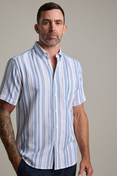 A man with short dark hair, a beard, and a tattooed right arm wears the Barkers Merten Stripe Shirt—light blue and white, short-sleeved, button-up—making it an ideal summer pick against a plain light background.