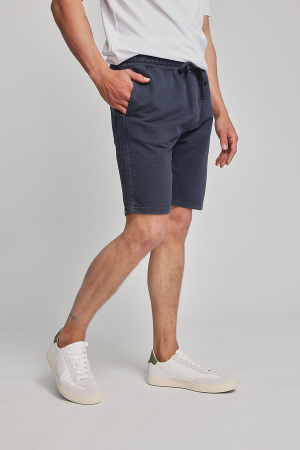 Someone wearing a white t-shirt, Barkers Organic Classic Print Track Short, and white sneakers stands with one hand in their pocket against a plain light background.