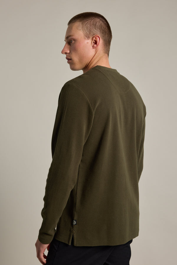 A young man with short hair wears the Barkers Avenue Waffle Henley in olive green and black pants, standing with his back facing slightly left and looking over his shoulder against a plain light background.