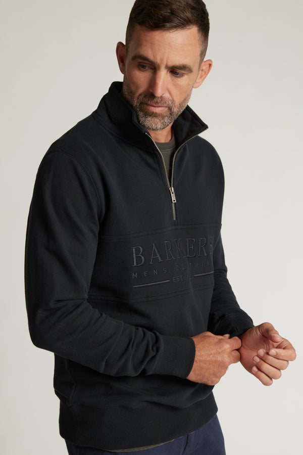 A man with short brown hair and a trimmed beard wears a black Barkers Heritage 1/4 Zip sweater, looking down as he adjusts his sleeve.