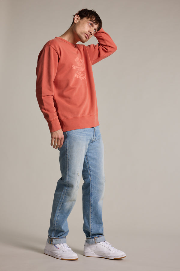 Wearing a coral Barkers Standard Issue Print Crew made from responsibly sourced cotton, light blue jeans, and white sneakers, a person stands against a plain background, tilting their head and touching the back of their head with one hand.
