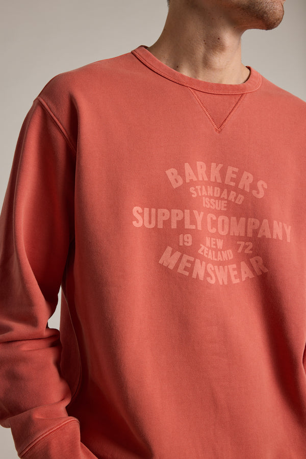 A person wears a faded red Barkers Standard Issue Print Crew, crafted from responsibly sourced cotton, with “Barkers Supply Company Menswear 1972 New Zealand” in matching red text. The face is partially out of frame.