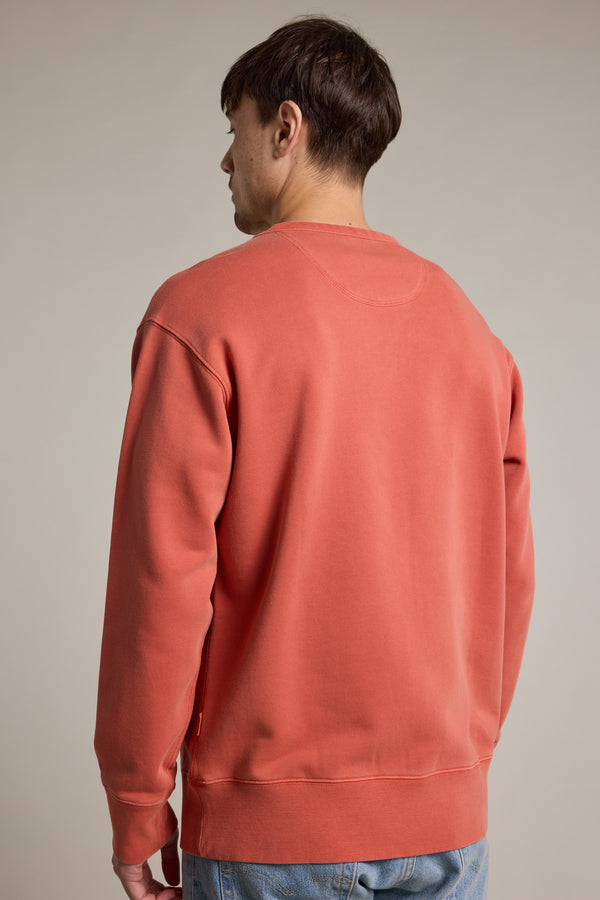 A man with short brown hair stands with his back to the camera, wearing a salmon Barkers Standard Issue Print Crew sweater made from responsibly sourced cotton loopback fleece, paired with light blue jeans against a plain neutral background.