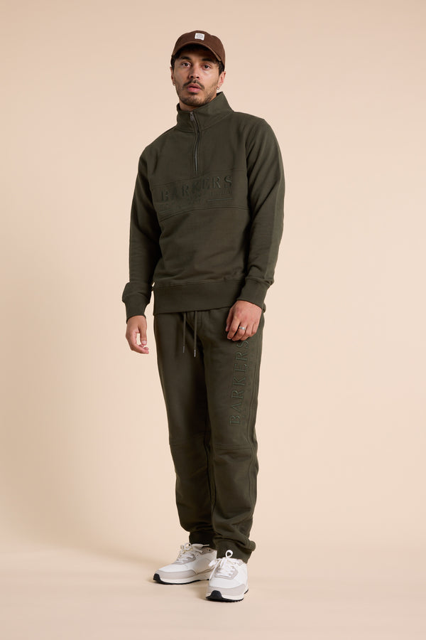 A man in an olive green Barkers Heritage 1/4 Zip by Barkers and matching sweatpants stands against a beige background, styled with a brown cap and white sneakers, gazing neutrally at the camera.