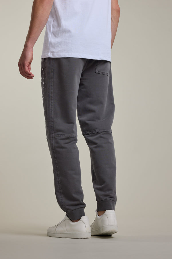 A person stands against a plain, light background wearing Barkers Organic Classic Print Track Pants with back patch pockets and ribbed cuffs, a white T-shirt, and white sneakers. The image is taken from behind, emphasizing the casual look.