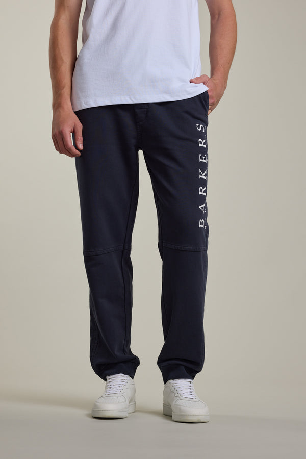 Someone stands against a plain background wearing a white tee, Barkers Organic Classic Print Track Pant with the logo vertically on the leg, and white sneakers.