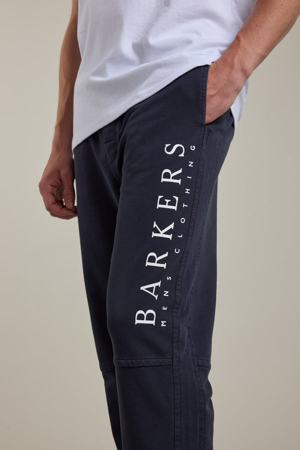 Someone wears Barkers Organic Classic Print Track Pant in dark blue with “BARKERS MENS CLOTHING” in white on the thigh and a white shirt, one hand in their pocket—classic ‘90s style updated for today.