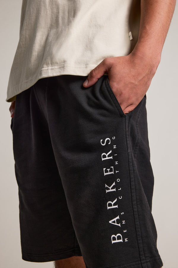A person wearing a light-colored T-shirt and relaxed fit Barkers Organic Classic Print Track Shorts, with one hand in their pocket. The black shorts display "BARKERS" printed vertically on the side.