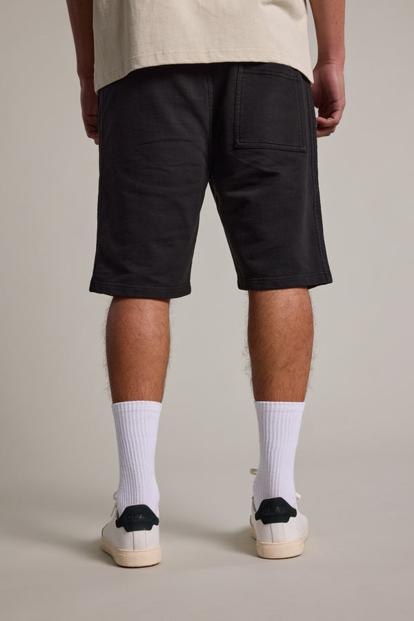 A person is shown from the back, wearing Barkers Organic Classic Print Track Shorts in black, white crew socks, and white sneakers with dark heels, standing against a plain light background.