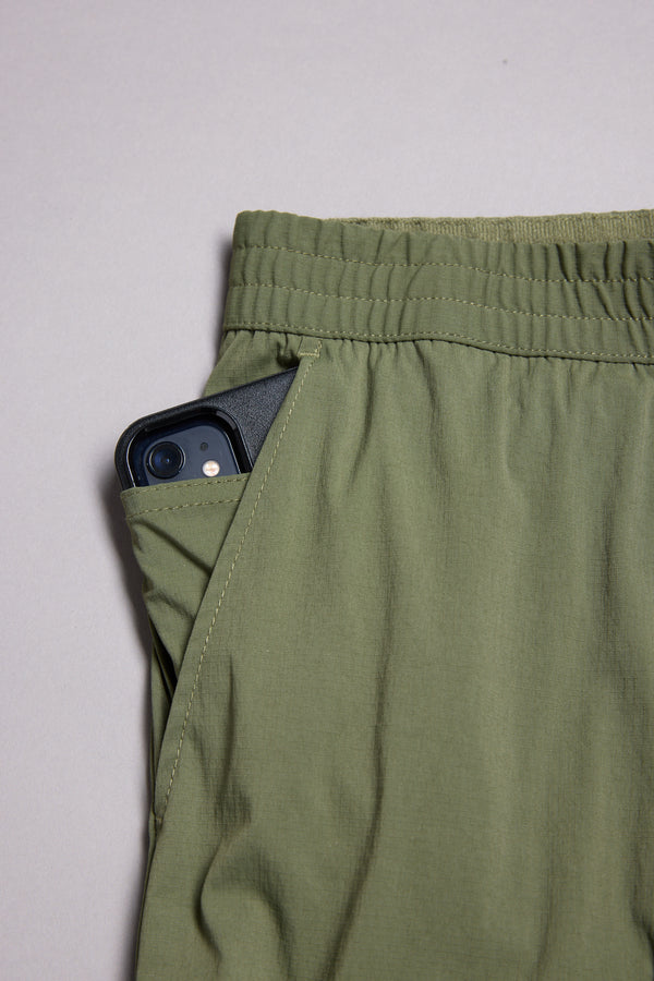 A black smartphone peeks from the side pocket of olive green WearEver Transit Pants by WearEver, featuring an elastic waist, shown against a neutral background.