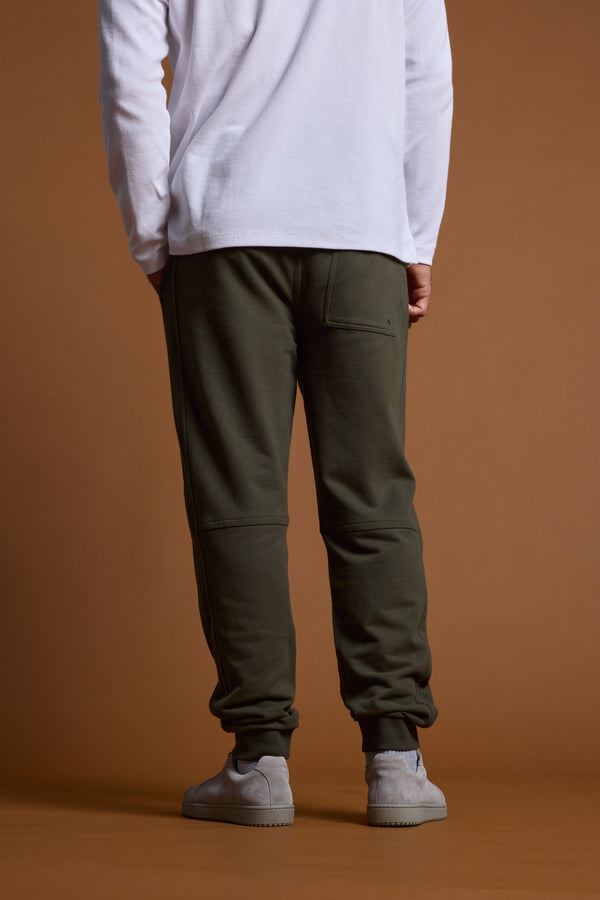 A person faces away, wearing a white long-sleeve shirt, Barkers Heritage Track Pant by Barkers in dark green with a back pocket, and light gray sneakers, set against a plain brown background.