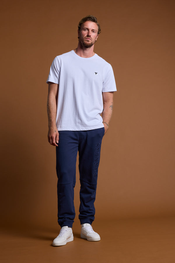 A man with short hair and a beard stands against a brown backdrop, wearing a white t-shirt, white sneakers, and navy Barkers Heritage Track Pants by Barkers, posing in a relaxed stance with one hand in his pocket.