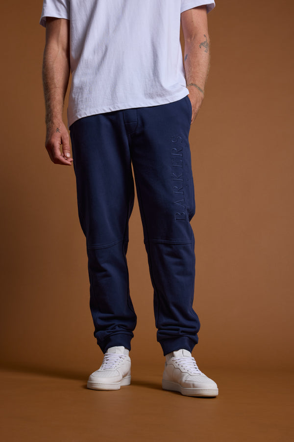 A person, shown from shoulders down, stands against a brown background wearing Barkers Heritage Track Pant in navy blue, paired with a white T-shirt and white sneakers.