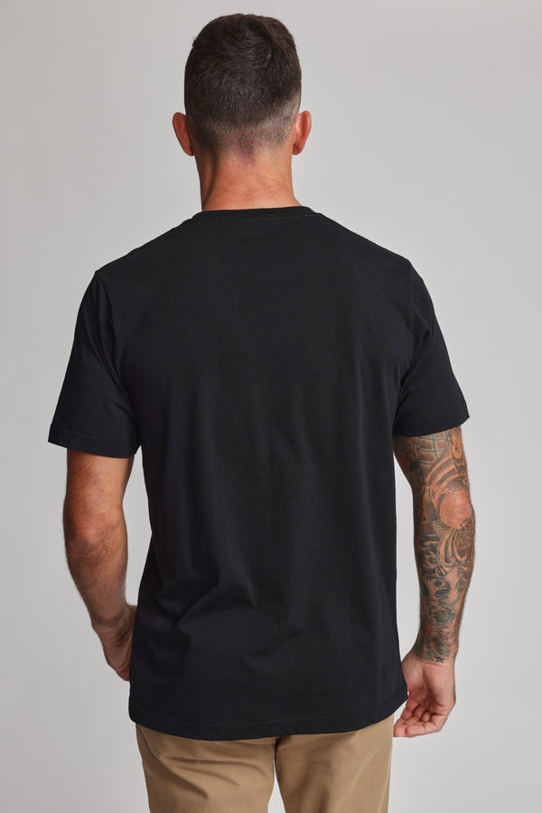 A man with short dark hair, a tattooed right arm, and tan pants stands with his back to the camera in a black Barkers Organic Crew Tee made from mid-weight organic cotton, set against a plain light background.