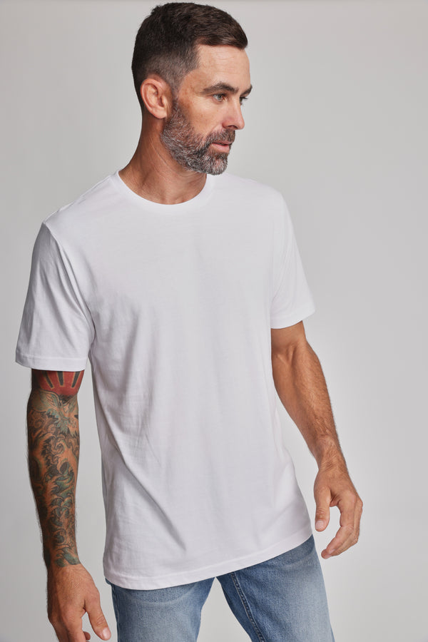 A man with short dark hair and a beard models the Barkers Organic Crew Tee in white with light blue jeans. He displays a colorful tattoo sleeve while standing against a plain light background, gazing to his left.