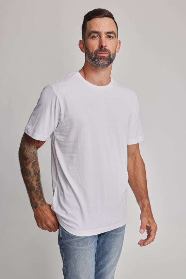 A man with short dark hair, a beard, and a tattooed right arm faces forward wearing the Barkers Organic Crew Tee in plain white with light blue jeans, standing against a light gray background.
