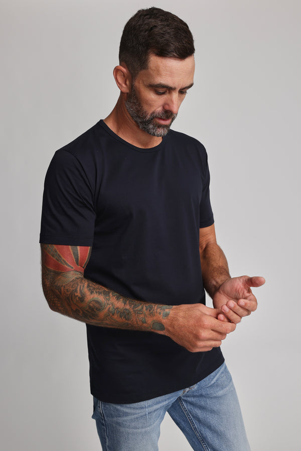 A man with short dark hair and a beard wears the Barkers Linton Supima Tee in plain black, paired with light blue jeans. He looks down, touching his wrist; colorful tattoos cover his left arm. The background is a plain light gray.