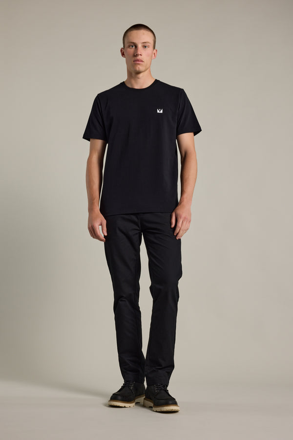 A man stands facing forward against a plain background, wearing the Barkers Newton Washed Tee—an oversized black box fit t-shirt with a small white logo on the chest—paired with black pants and black shoes.