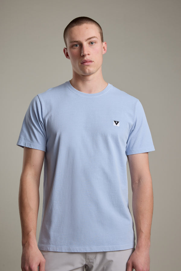 A young man with short hair wears the Barkers Newton Washed Tee, a light blue organic cotton shirt featuring a small black and white logo on the left chest, standing against a plain light gray background.