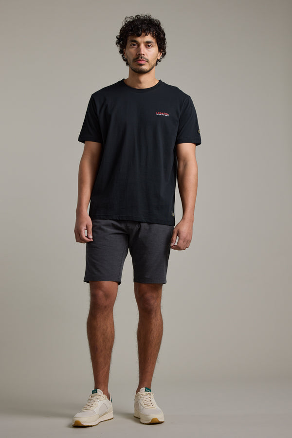 A man with curly hair stands against a plain background, wearing a Legasea Brand Tee by Legasea. Facing forward with a neutral expression and relaxed arms, he embodies support for New Zealand marine conservation.