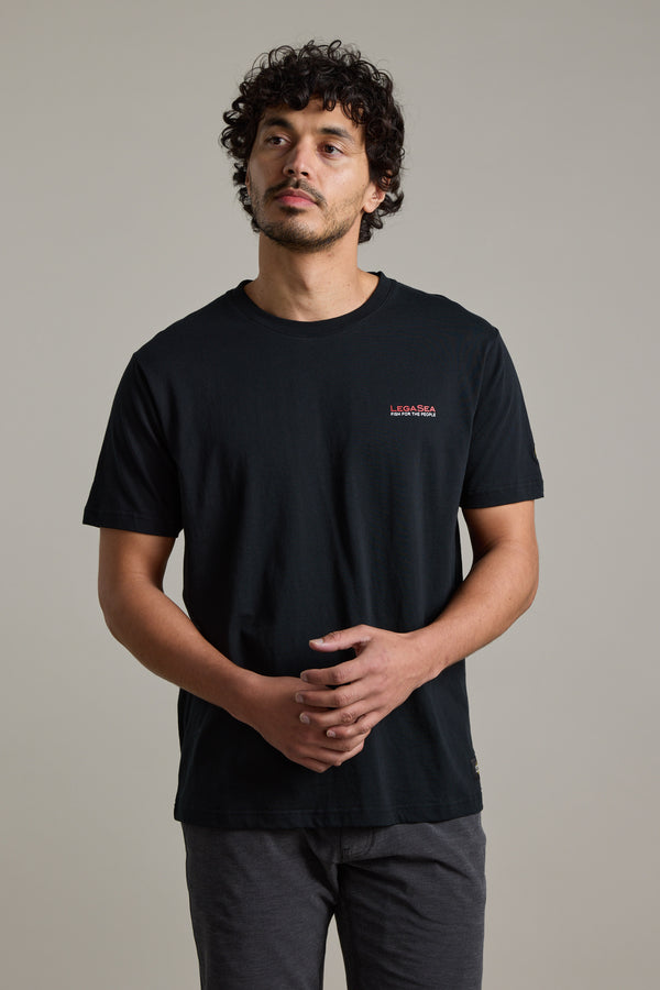 A man with curly dark hair and facial hair stands against a plain background, wearing the Legasea Brand Tee by Legasea. Made from organic cotton, the tee reflects New Zealand marine conservation values as he looks upward, hands loosely clasped.