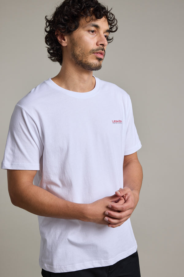A man with curly hair and light facial hair wears the Legasea Brand Tee by Legasea and black pants, standing against a neutral background with his hands lightly clasped in front of him.