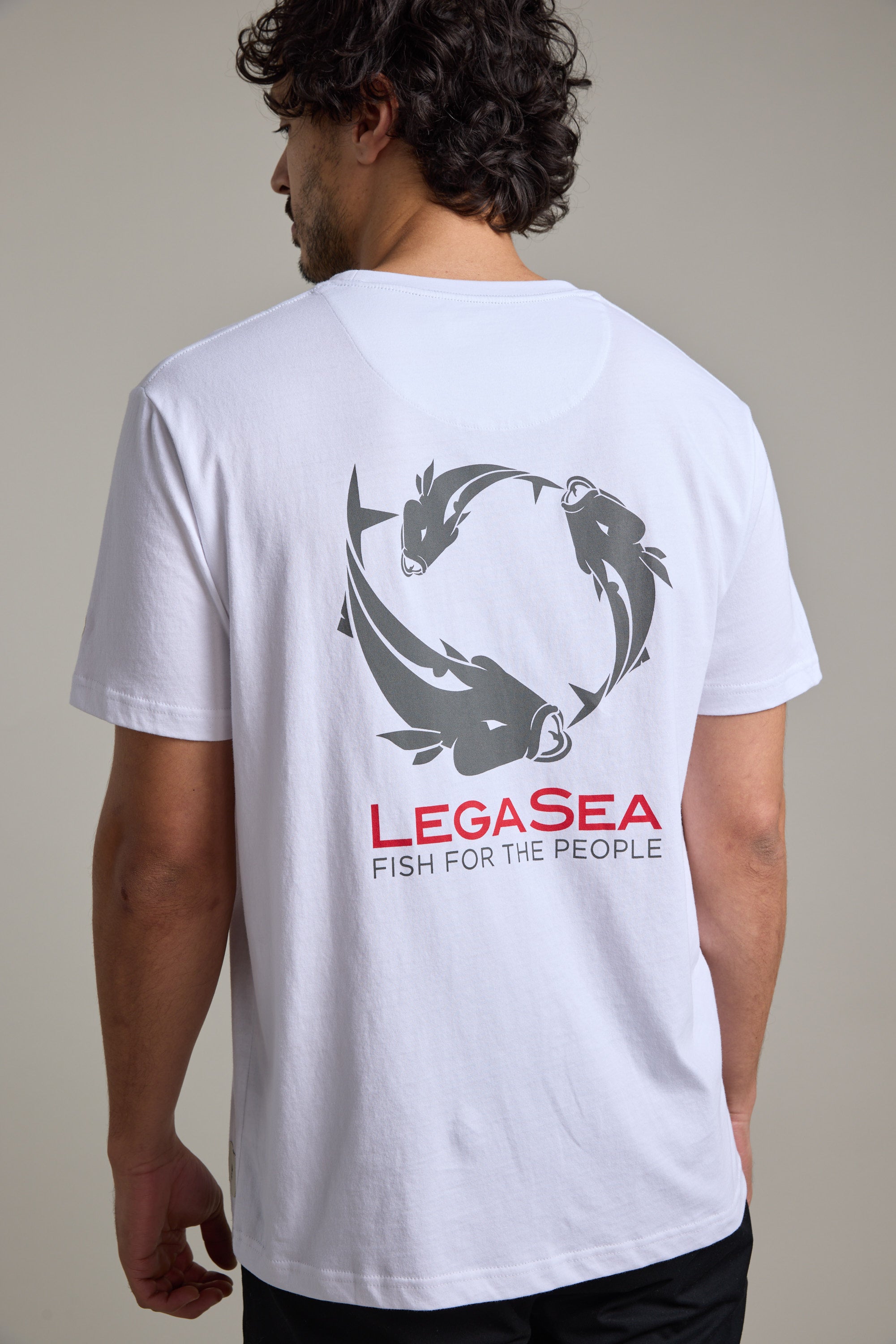 A man wears the Legasea Brand Tee, made from organic cotton, with a circular fish graphic and "LEGASea FISH FOR THE PEOPLE" in red and gray on the back as he faces away, clearly displaying the Legasea design.