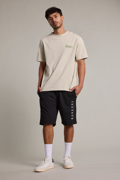 A man stands against a plain background wearing a beige Barkers T-shirt, black Barkers Organic Classic Print Track Shorts, white crew socks, and white sneakers, hands in pockets and looking slightly upward.