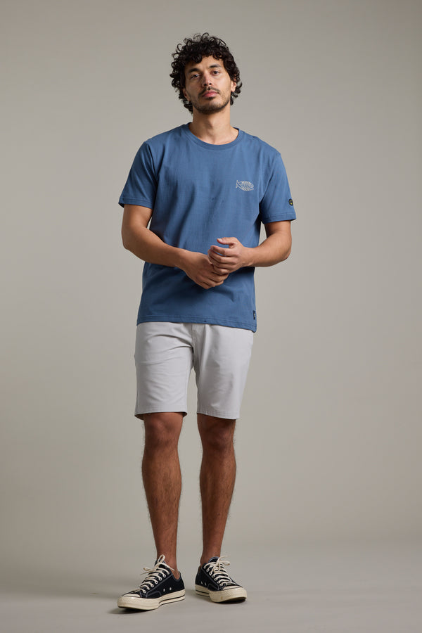 A man with curly hair faces forward, wearing a blue T-shirt, Barkers Hutchinson Walkshort in light gray, and black sneakers with white soles, set against a plain light gray background.