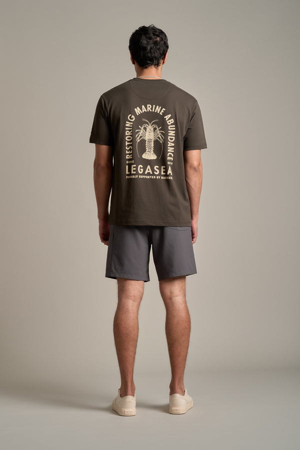 A man faces away, wearing the Legasea Crayfish Print Tee—an organic cotton t-shirt from Legasea with marine-themed back graphics—paired with gray shorts and light shoes. Supports New Zealand marine conservation.