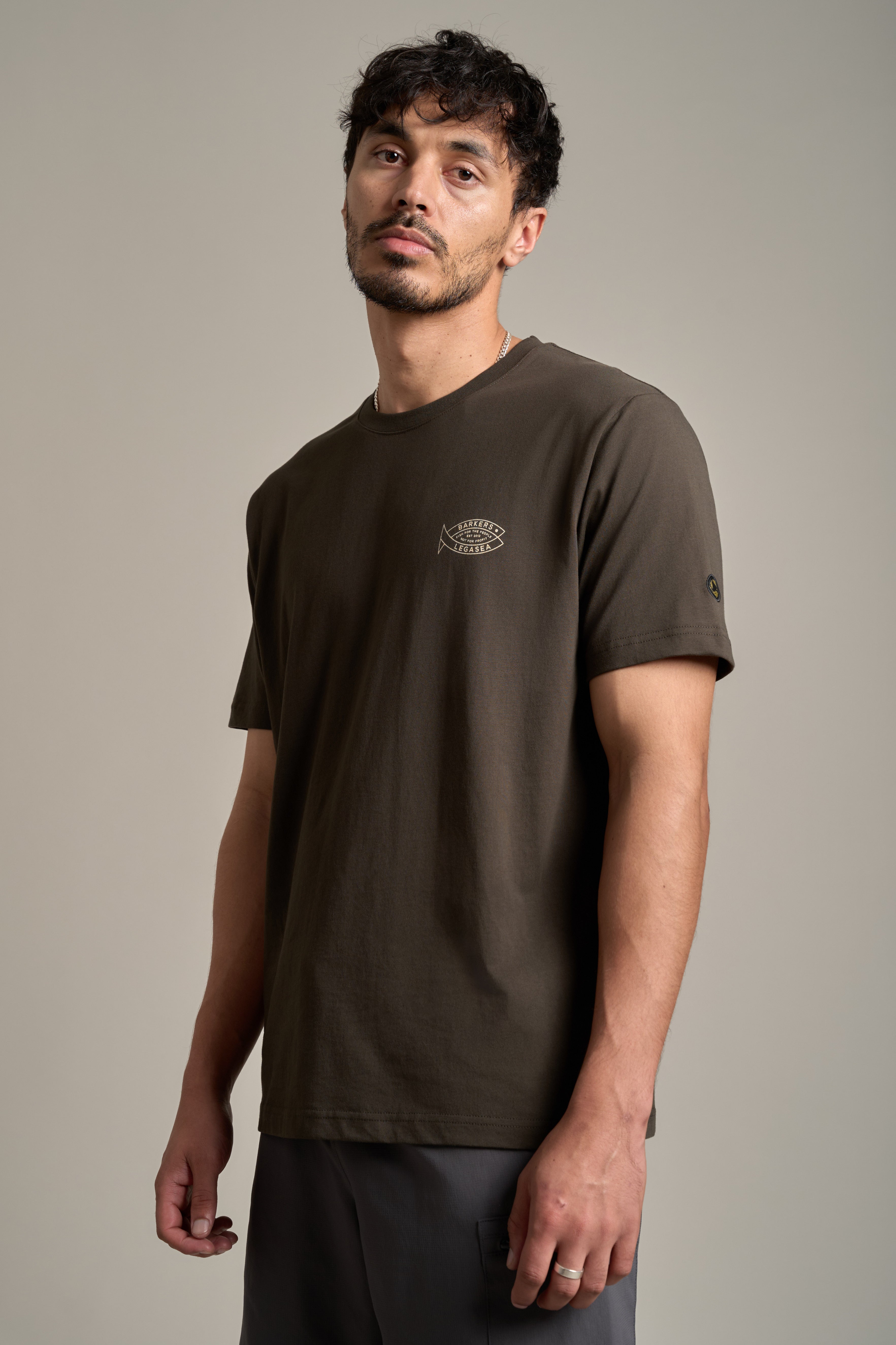 A man with short curly hair and facial hair wears a dark olive Legasea Crayfish Print Tee, made from organic cotton, featuring a small fish graphic on the chest and dark pants. Inspired by New Zealand marine conservation, he stands against a plain background.