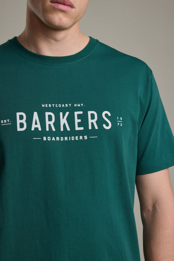A person wears the Barkers Boardriders Logo Tee in dark green organic cotton, featuring the Westcoast Hwy. logo and "Est. 1972" on the front. The photo highlights this everyday essential by focusing on the upper torso and part of their face.